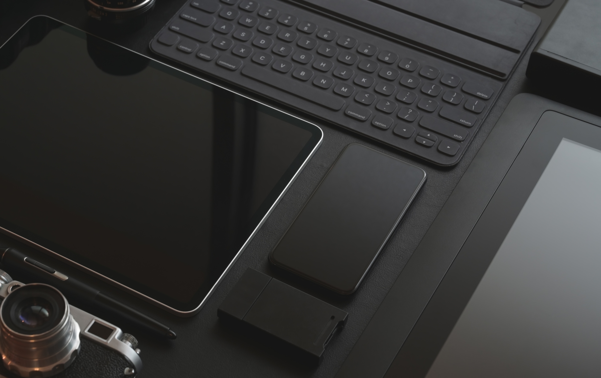 黑色極簡風工作桌面，擺放平板電腦、智慧型手機、鍵盤與相機配件，呈現科技感與專業質感的設計場景。