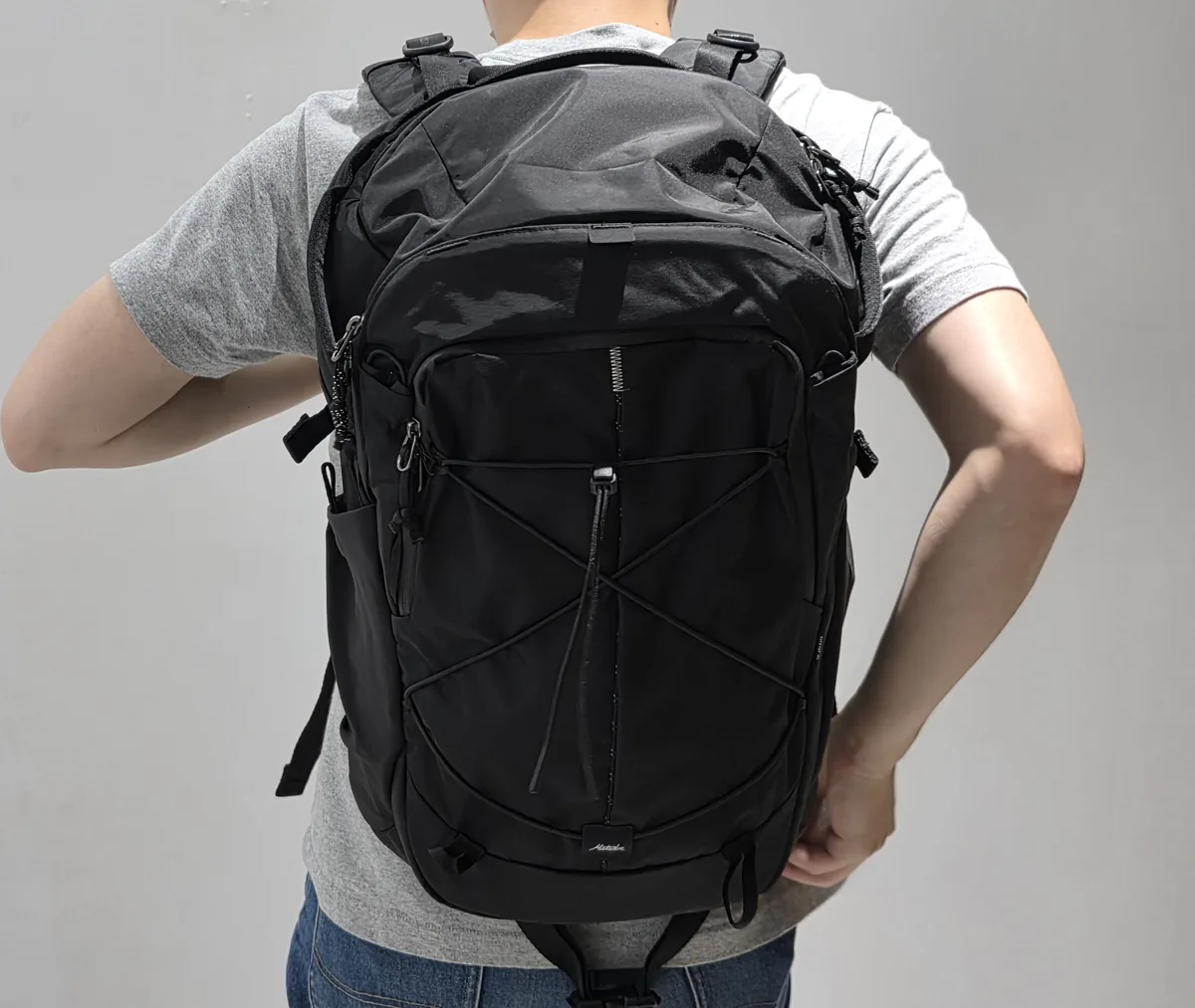 使用者背著黑色 Adventure Pack 24L 的後視畫面，前袋交叉彈性繩與兩側水壺袋朝外