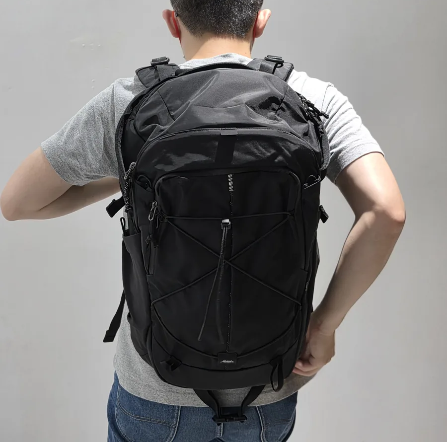 使用者背著黑色 Adventure Pack 24L 的後視畫面，前袋交叉彈性繩與兩側水壺袋朝外